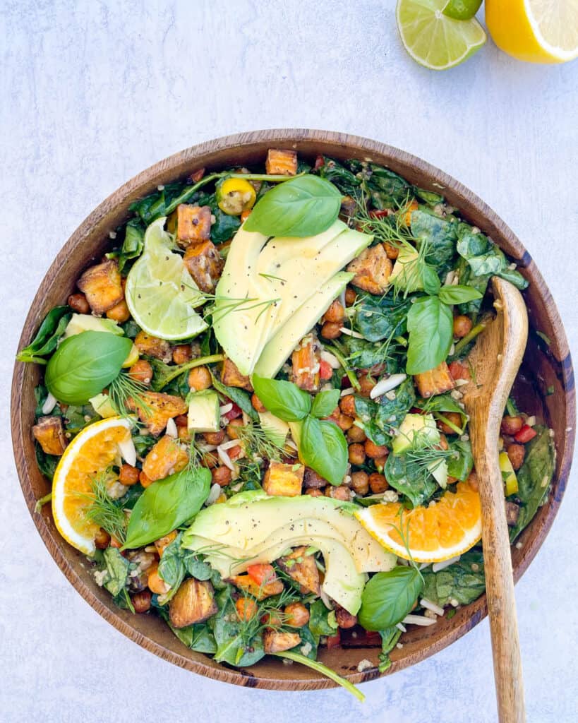 salad in a wooden bowl topped with avocado sliced orange sliced lime basil leaves chickpeas and baby spinach with a wooden spoon in the bowl slices limes are placed in the top right corner next to the bowl