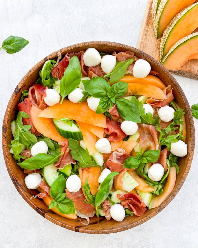 cantaloupe salad with arugula prosciutto mozzarella balls fresh basil and cucumber in a wooden bowl with a wooden spoon in the bowl. sliced of cantaloupe on cutting board shown in the top right corner