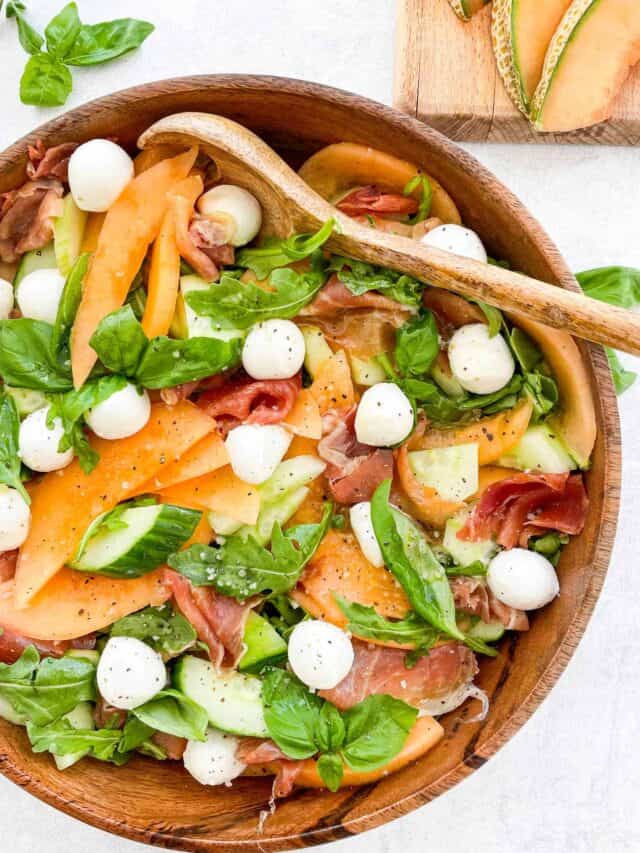 cantaloupe salad with arugula prosciutto mozzarella balls fresh basil and cucumber in a wooden bowl with a wooden spoon in the bowl. slices of cantaloupe on a wooden cutting board in top right corner