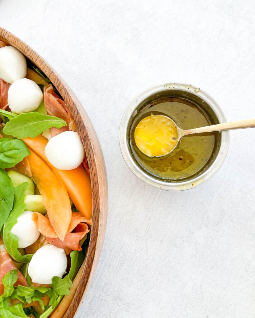 close up homemade vinaigrette in a small metal ramekin with a gold spoon inside. on the left is a cut off bowl of the cantaloupe salad with mozzarella balls prosciutto and basil