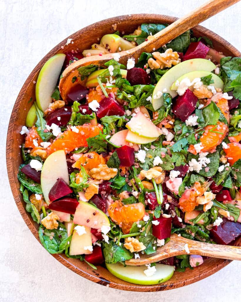 overhead view of beet salad with feta cheese crumbles, sliced apples, orange slices, arugula, spinach, and chopped walnuts in a wooden bowl with two large wooden spoons inside the bowl