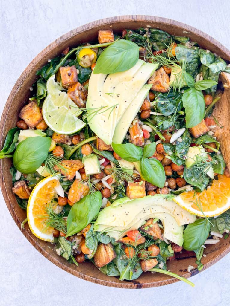 salad in a wooden bowl topped with avocado sliced orange sliced lime basil leaves chickpeas and baby spinach with a wooden spoon in the bowl 