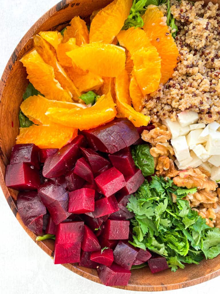 close up of sliced beets, cilantro, chopped walnuts, feta cheese, orange slices, and quinoa in a wooden bowl