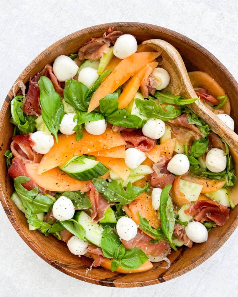 close up of cantaloupe salad with arugula prosciutto mozzarella balls fresh basil and cucumber in a wooden bowl with a wooden spoon in the bowl