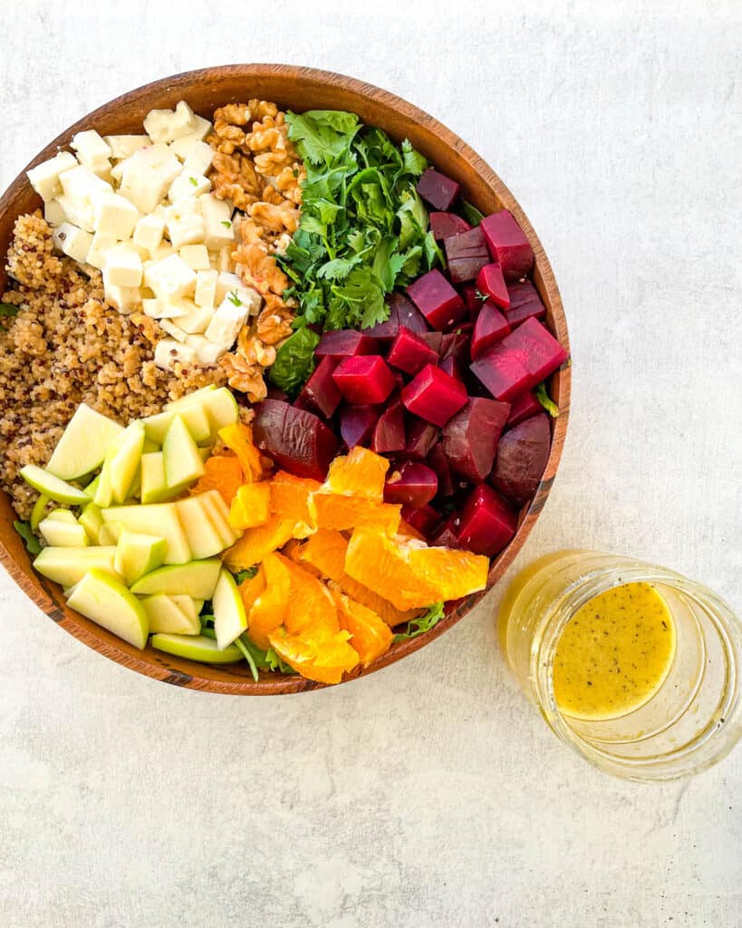 overhead view of beet salad with feta cheese crumbles, sliced apples, orange slices, arugula, spinach, cilantro, and chopped walnuts in a wooden bowl with a mason jar of orange vinaigrette dressing on the side of the bowl