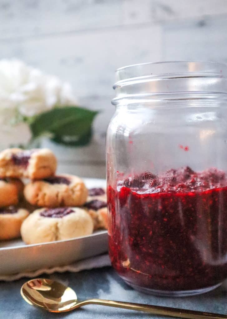 mason jar filled with homemade raspberry jam and a gold spoon in front of the jar and a plate of thumbprint cookies in the background slightly blurred