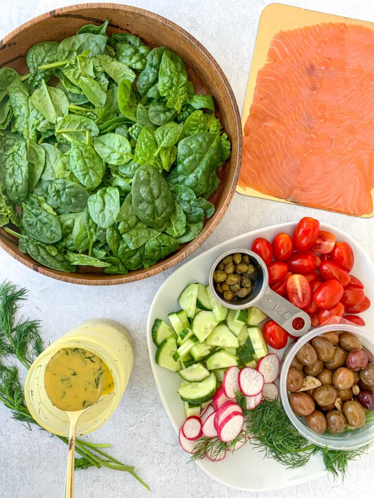 ingredients needed shown are smoked salmon baby spinach in a large wooden bowl honey dijon dill vinaigrette in a small mason jar with gold spoon and a plate of assorted vegetables that include olives capers tomatoes cucumbers radish and dill
