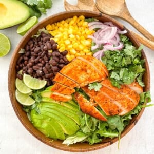 southwest chicken salad in a large wooden bowl with two wooden spoons next to the bowl and a half avocado and limes in the top left corner