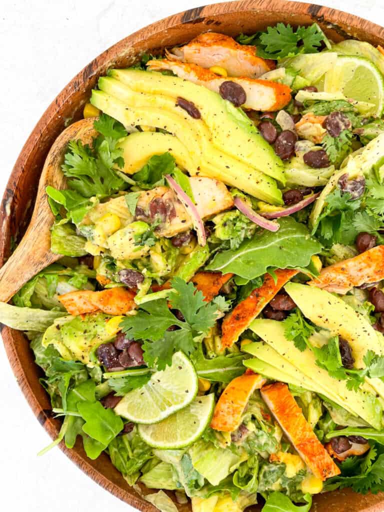 grilled chicken cilantro onions corn black beans sliced avvocado and lime wedges in a bowl assembled to make a southwest chicken salad two wooden spoons on top right next to the bowl and on top left an sliced avocado and limes next to the bowl