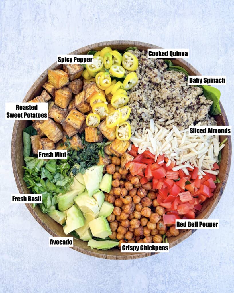 ingredients to make superfood salad shown in a large wooden bowl are roasted sweet potatoes sliced yellow spicy peppers cooked quinoa sliced almonds crispy chickpeas sliced avocado mint basil all on a bed of baby spinach