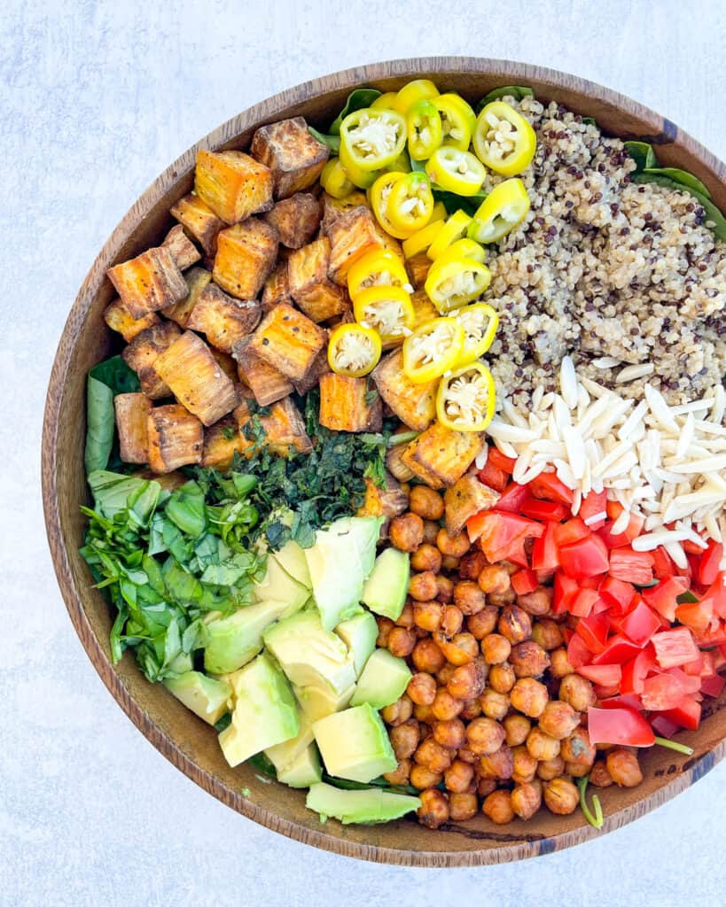 ingredients to make superfood salad shown in a large wooden bowl are roasted sweet potatoes sliced yellow spicy peppers cooked quinoa sliced almonds crispy chickpeas sliced avocado mint basil all on a bed of baby spinach