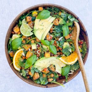 salad in a wooden bowl topped with avocado sliced orange sliced lime basil leaves chickpeas and baby spinach with a wooden spoon in the bowl