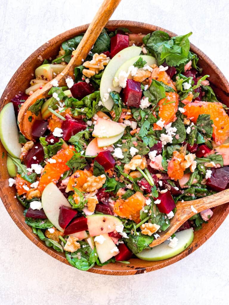 overhead view of beetroot salad with feta cheese crumbles, sliced apples, orange slices, arugula, spinach, and chopped walnuts in a wooden bowl with two large wooden spoons inside the bowl