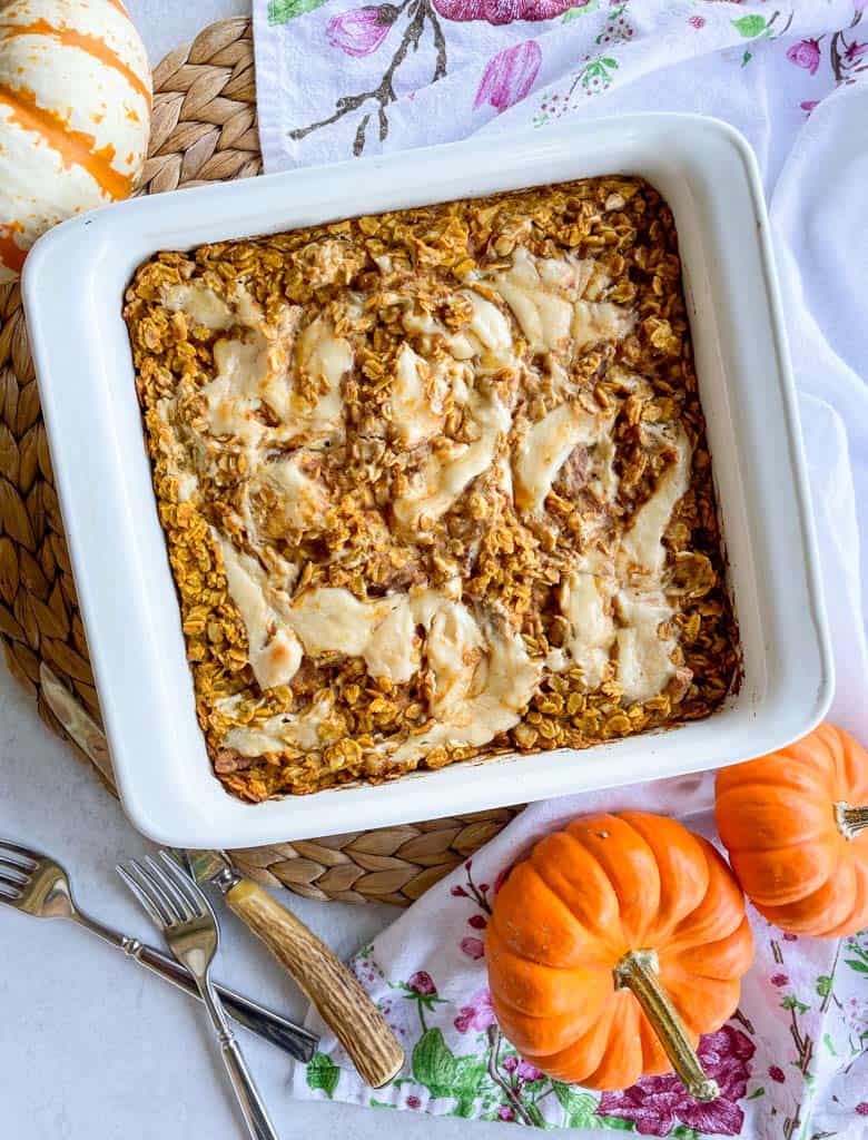 pumpkin baked oatmeal with maple cream cheese swirl in a white baking dish with small pumpkins in the corners of the photo and utensils next to the baking dish