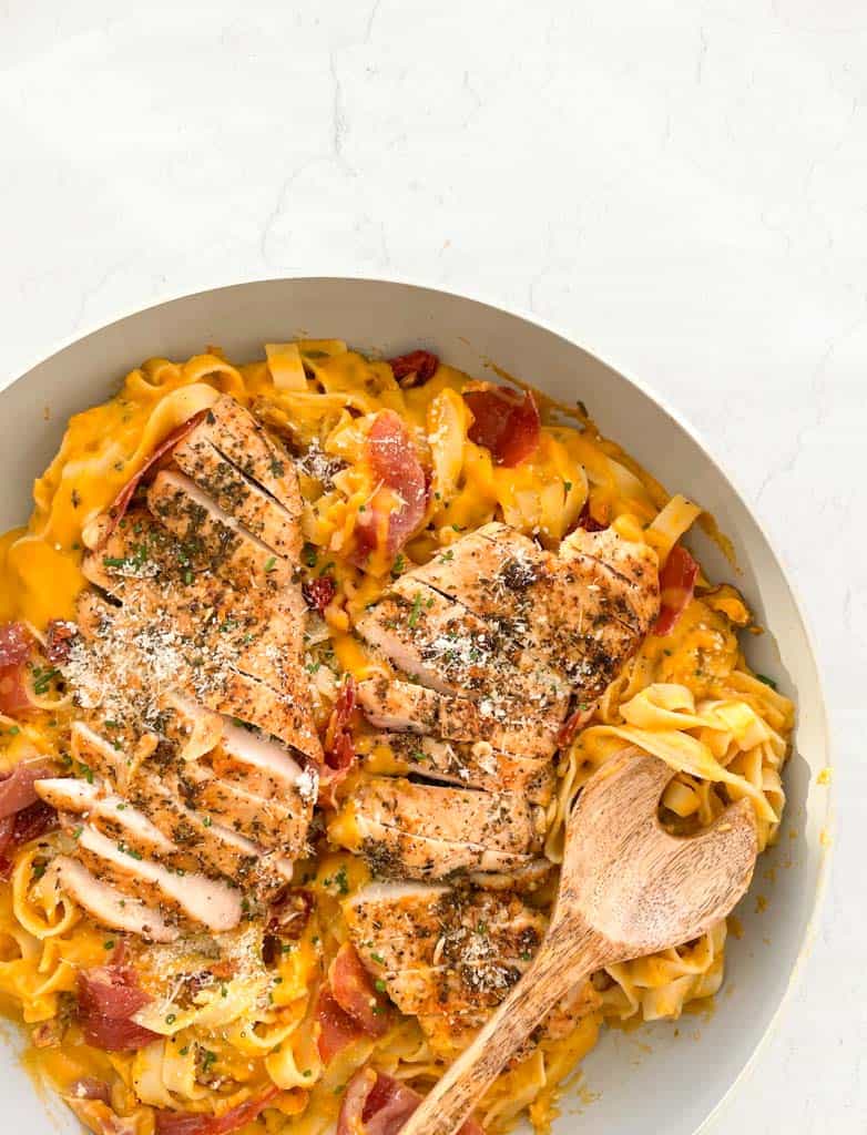 butternut squash pasta topped with crispy prosciutto and sauteed chicken and parmesan cheese in a skillet with a wooden spoon