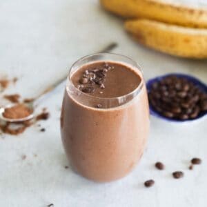 coffee smoothie in a glass with a spoon of cacao powder in the background and two banana and a small bowl of coffee beans