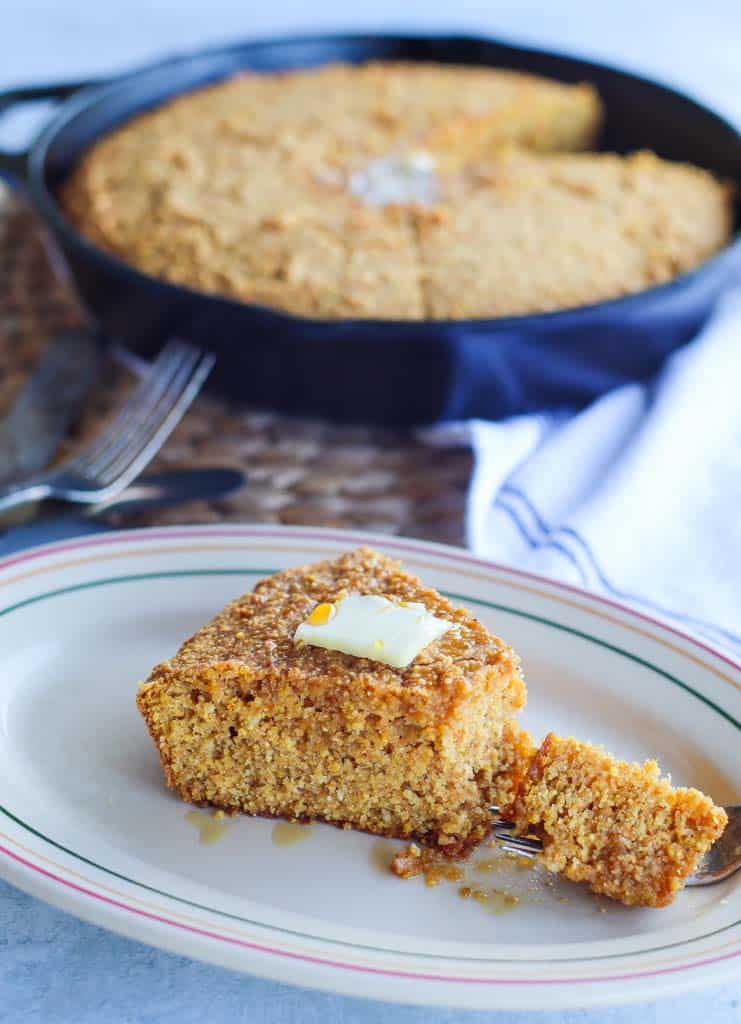 A slice of sweet potato cornbread on a plate with butter on top and a skillet full of cornbread in the background