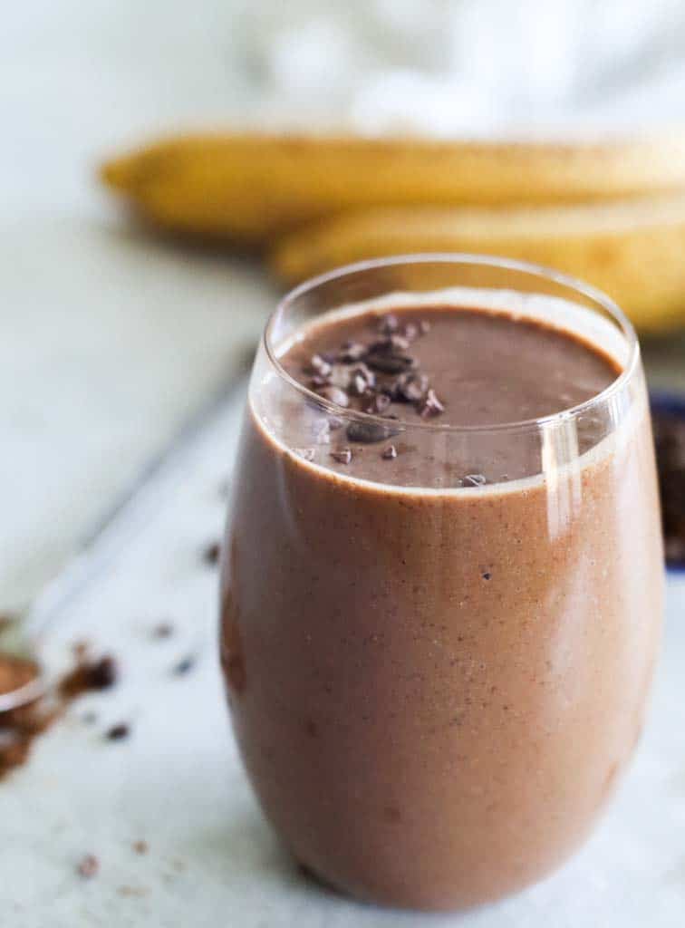 coffee smoothie in a glass with a spoon of cacao powder in the background and two banana and a small bowl of coffee beans