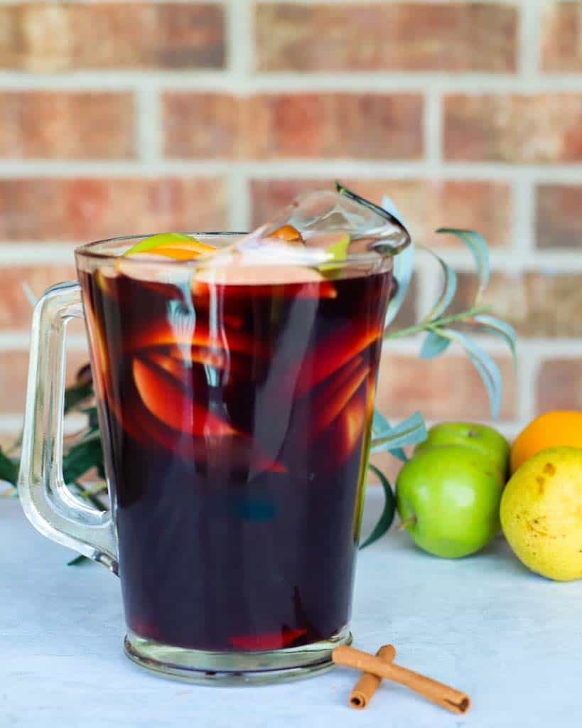 a pitcher of red fall sangria with seasonal fruits in the background and two cinnamon sticks