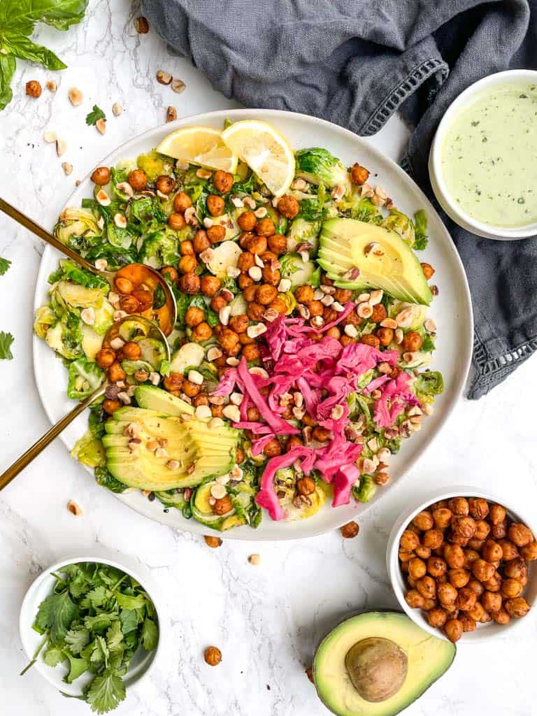 warm brussel sprout salad on a white plate with two gold spoons a ramekin of creamy vinaigrette is in the top right cover a small bowl of chickpeas in the the bottom right side of the photo next to a half avocado on the bottom left is a ramekin of cilantro