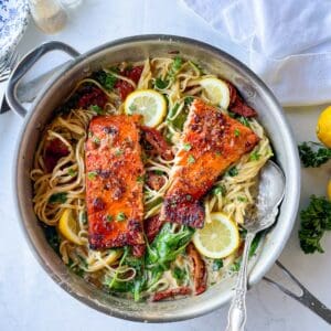 two salmon fillets in a large skillet filled with pasta in a creamy dairy free sauce. The pasta is garnished with fresh parsley and lemon wedges. a serving spoon is in the skillet