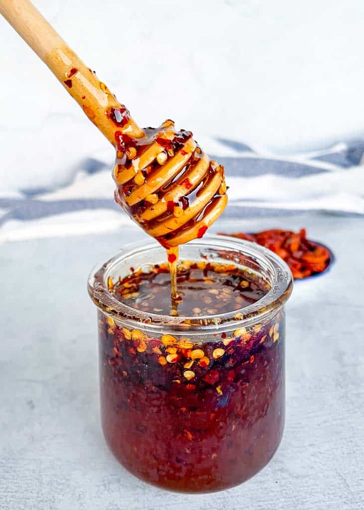 Hot honey sauce in a small glass jar with a honey comb spoon covered in honey and red pepper flakes being held over the jar