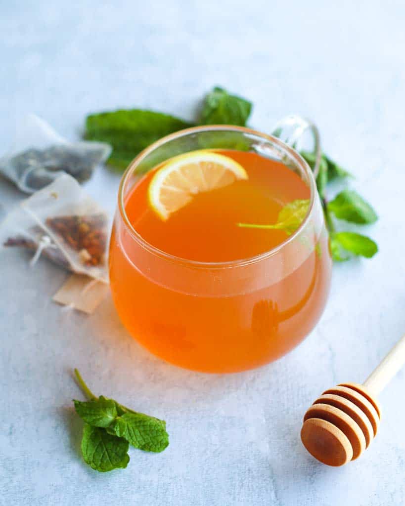 a cup of mint tea surrounded by mint leaves, tea bags and a honeycomb stick