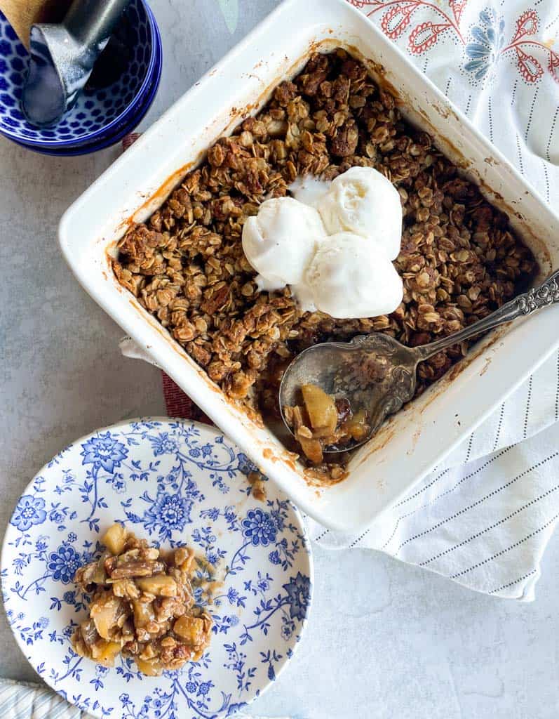 A dish of apple crisp topped with vanilla ice cream with a plate of a smaller portion on the side.