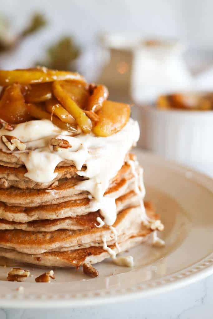 a stack of apple cinnamon pancakes on a white plate topped with maple cream cheese glaze chopped pecans and caramelized apple slices 