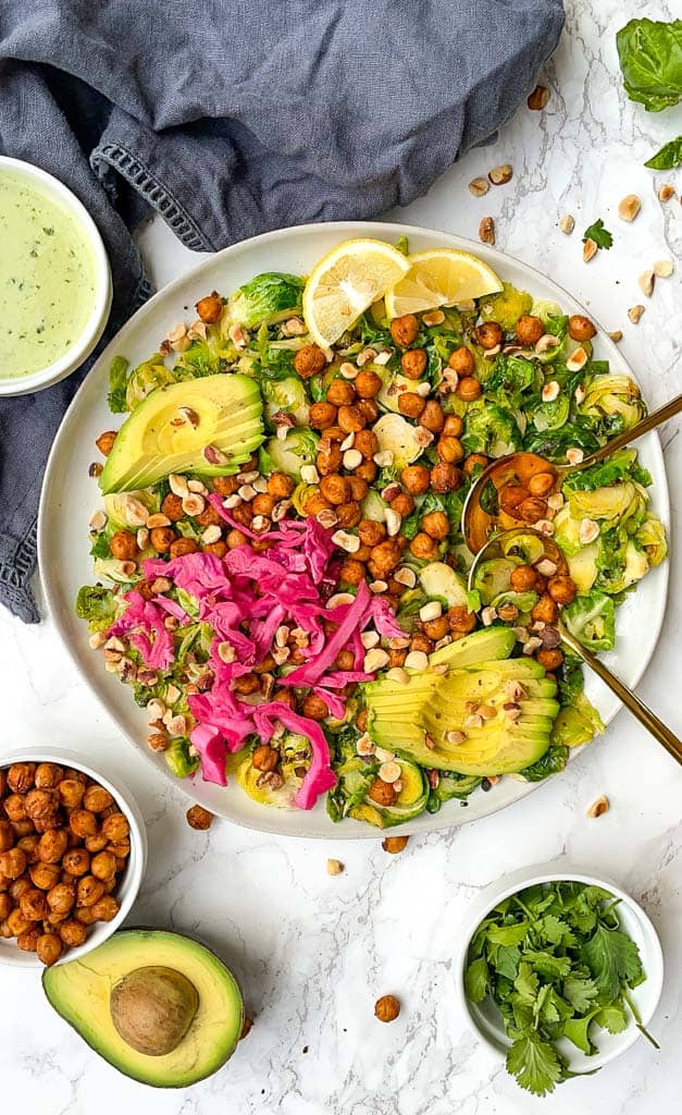 warm brussel sprout salad on a white plate with two gold spoons a ramekin of creamy vinaigrette is in the top left is a small bowl of chickpeas in the the bottom left side of the photo next to a half avocado on the bottom right is a ramekin of cilantro
