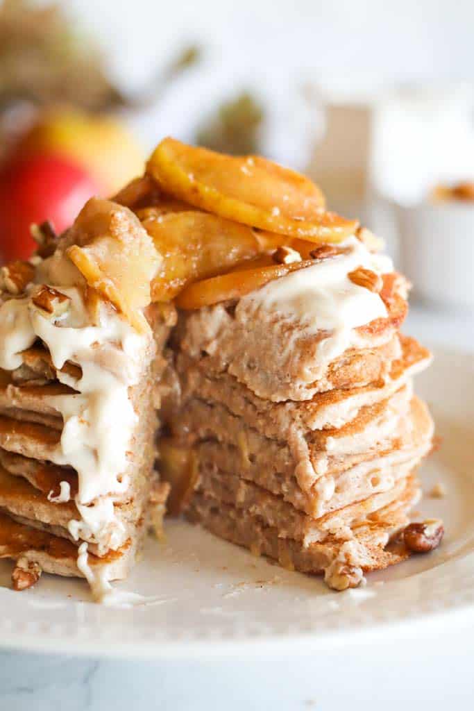 a stack of apple cinnamon pancakes on a white plate topped with maple cream cheese glaze chopped pecans and caramelized apples with a large portion sliced to show inner part of the pancakes