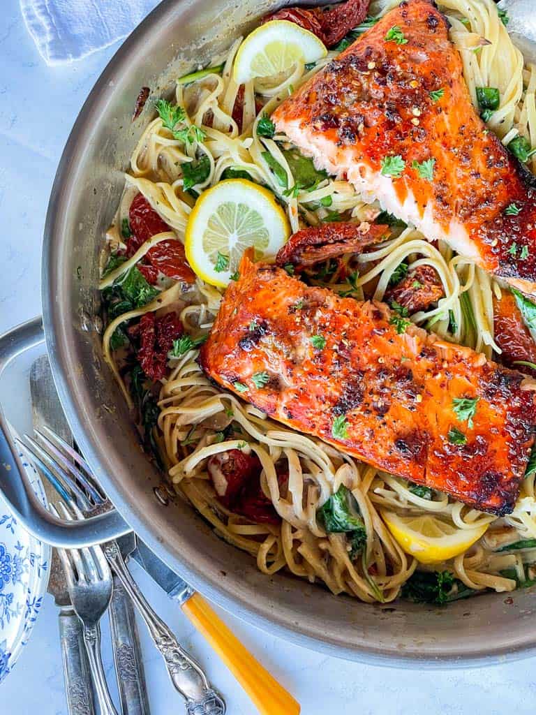 two salmon fillets in a large skillet filled with pasta in a creamy dairy free sauce. The pasta is garnished with fresh parsley and lemon wedges. forks and knives are on the side of the skillet