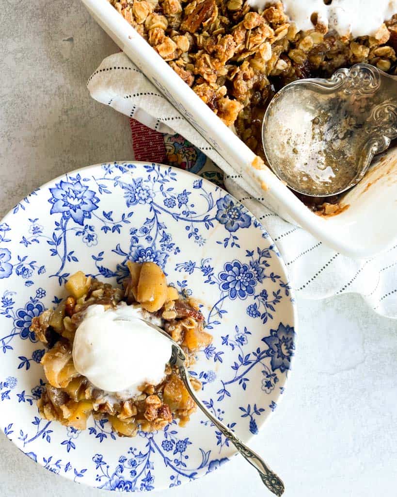 a single serving of apple crisp topped with a scoop of ice cream with a pan of the dessert off to the side
