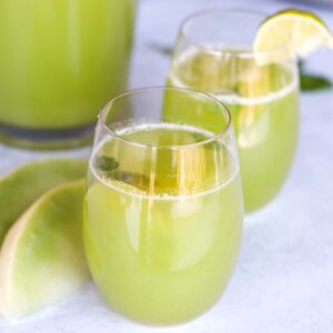 Honeydew juice in two glasses with a pitcher of honeydew juice in the background in the right corner and two slices of honeydew melon next to the glasses