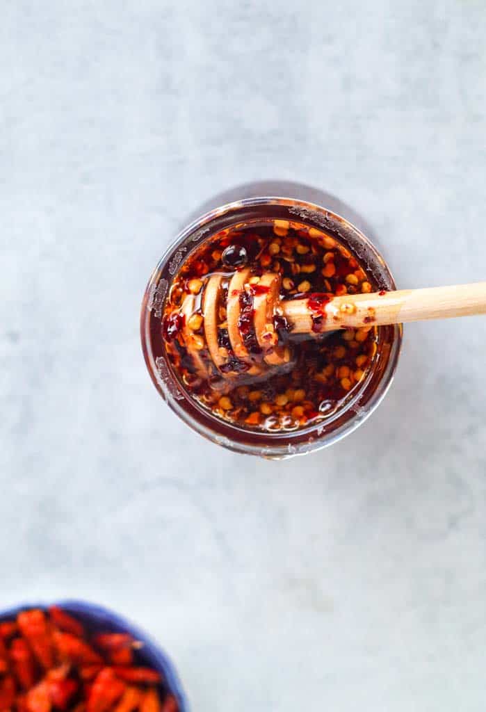 Hot honey sauce in a small glass jar with a honey comb spoon covered in honey and red pepper flakes being held over the jar and a small bowl of chile peppers in the bottom left corner