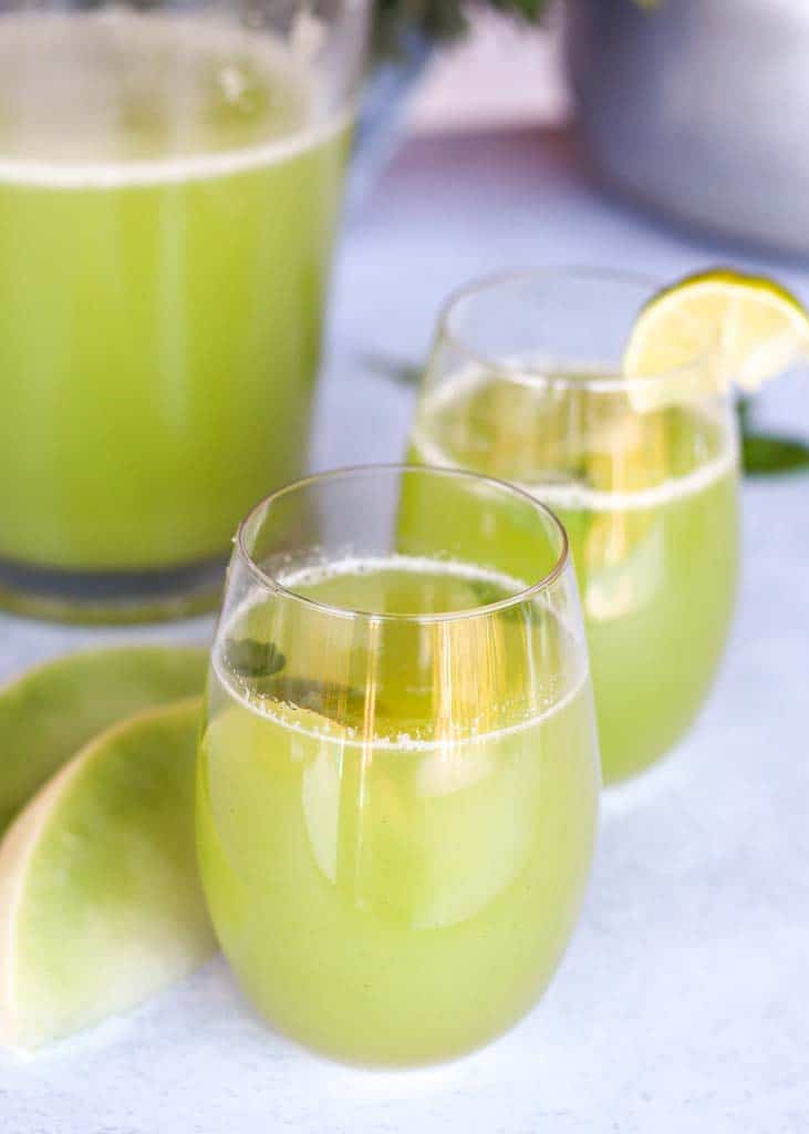 Honeydew juice in two glasses with a pitcher of honeydew juice in the background in the right corner and two slices of honeydew melon next to the glasses