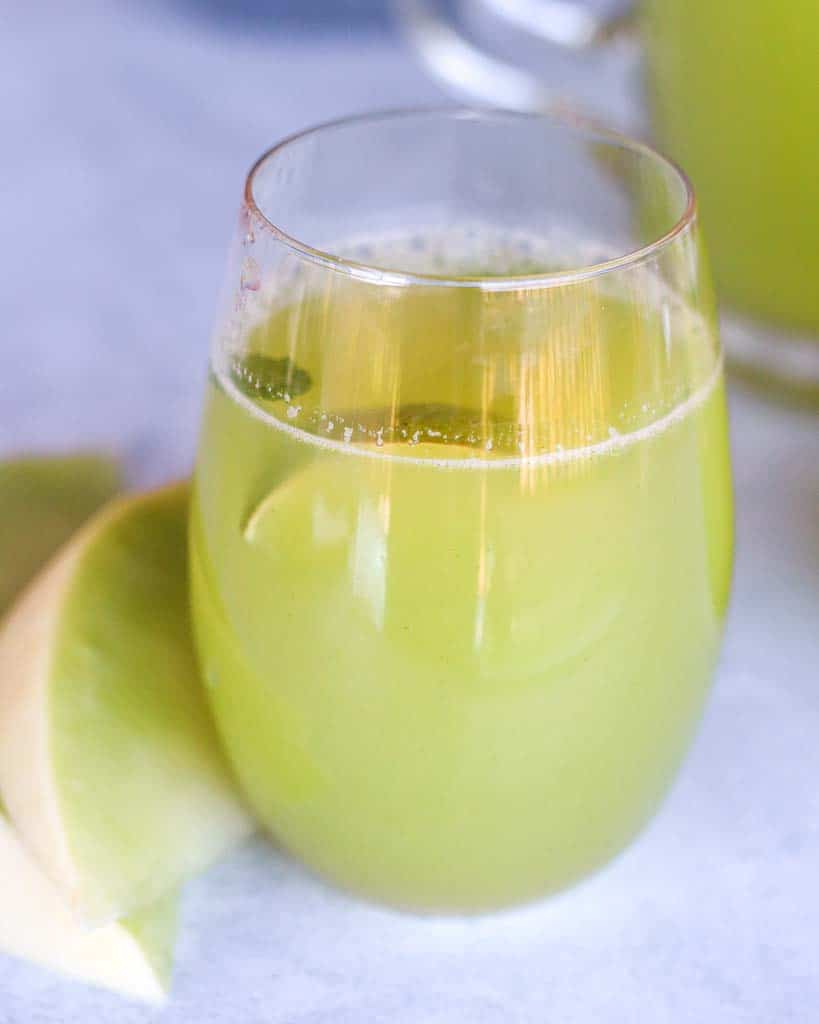one glass of honeydew melon juice with two slices of honeydew to the right of the juice and a pitcher of honeydew in the top left corner