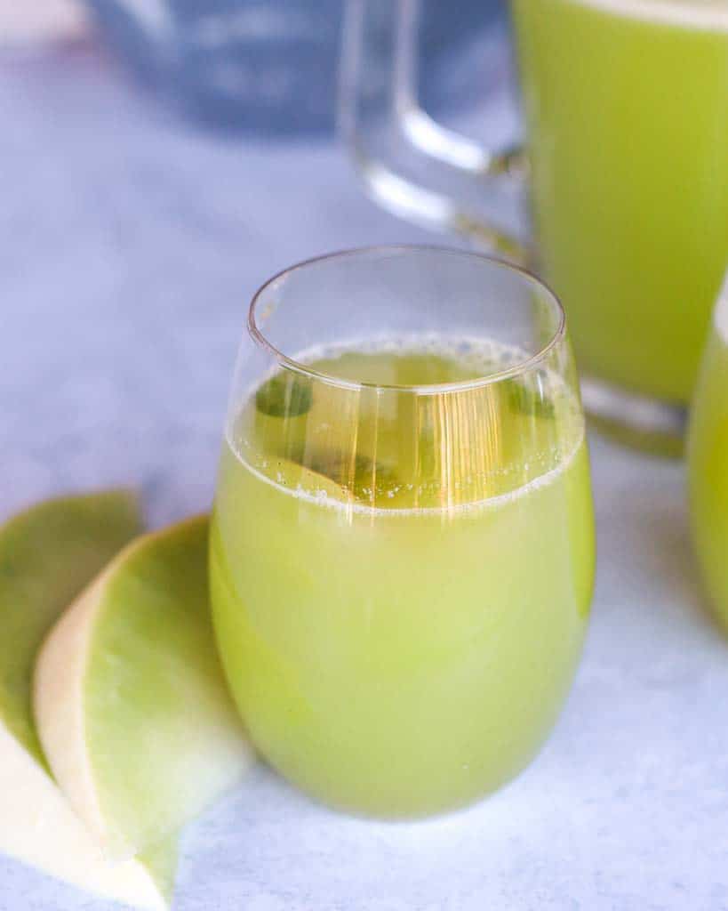 one glass of honeydew melon juice with two slices of honeydew to the right of the juice and a pitcher of honeydew  in the top left corner
