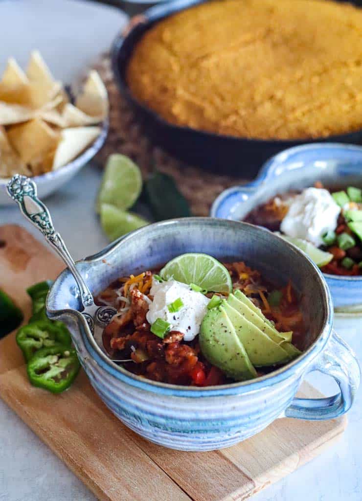 a spoon in a bowl of chili with tortilla chips and a skillet of cornbread in the background