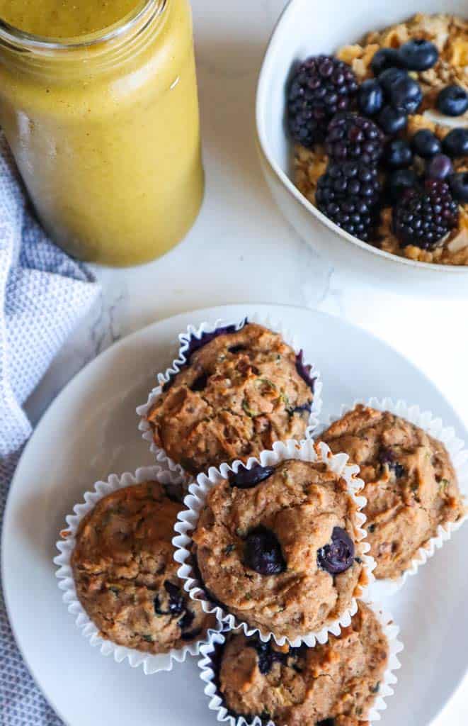 a plate of zucchini blueberry muffins with a yellow smoothie and a bowl of berries and granola in the top right corner