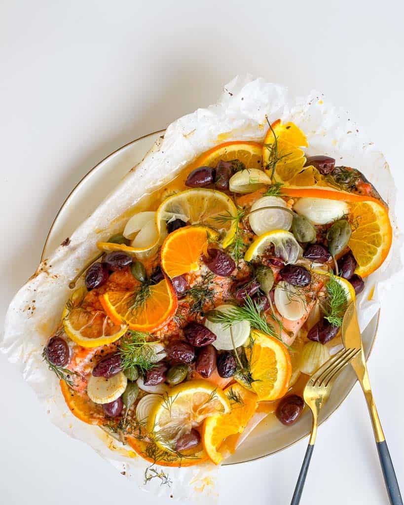 baked rainbow trout with slices lemon oranges kalamata olives fresh dill capers on parchment paper on a white plate with a fork and knife