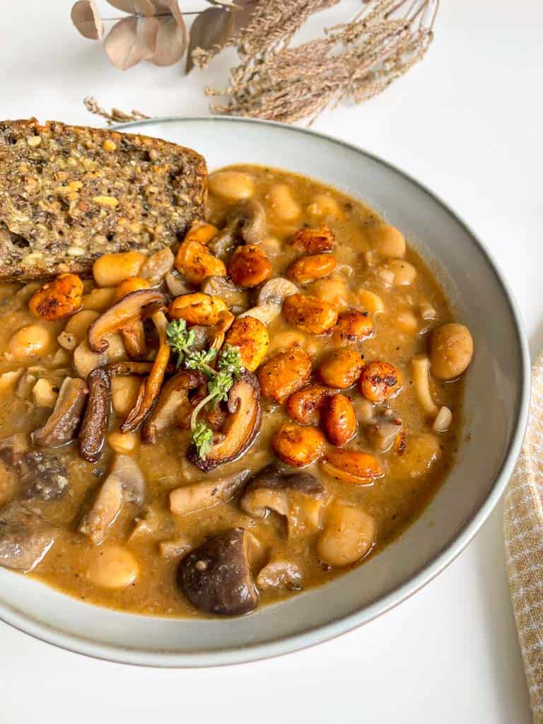 White bean and mushroom soup topped with crispy white beans and thyme in a white bowl with a piece of gluten free seed bread