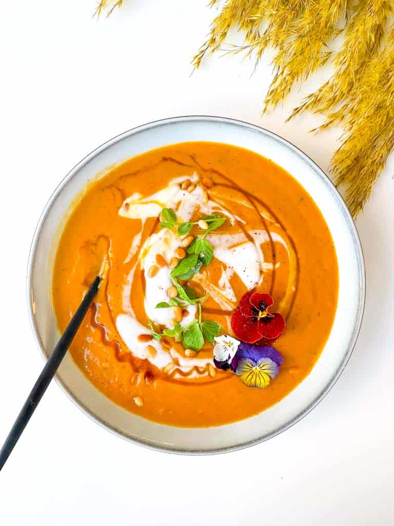 roasted butternut squash soup in a white bowl topped with a dollop of yogurt honey micro greens edible flowers and toasted pine nuts with a spoon in the bowl