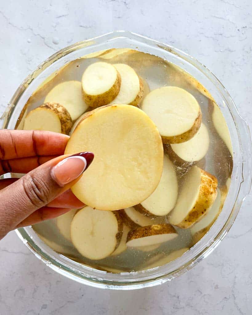 a bowl of sliced potatoes submerged in water with a hand holding one slice