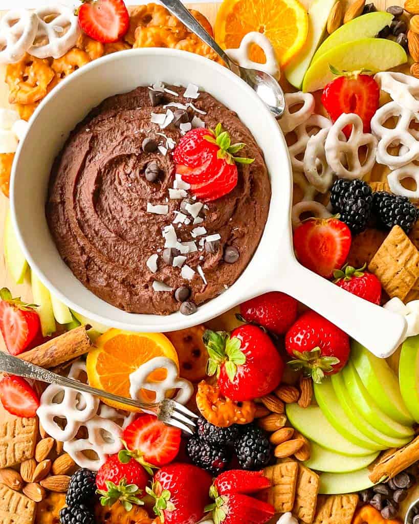 chocolate hummus in a white dish surrounded by pretzels orange slices strawberries sliced apples blackberries and crackers for dipping into the hummus