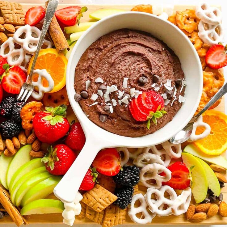 chocolate hummus in a white dish surrounded by pretzels orange slices strawberries sliced apples blackberries and crackers for dipping into the hummus