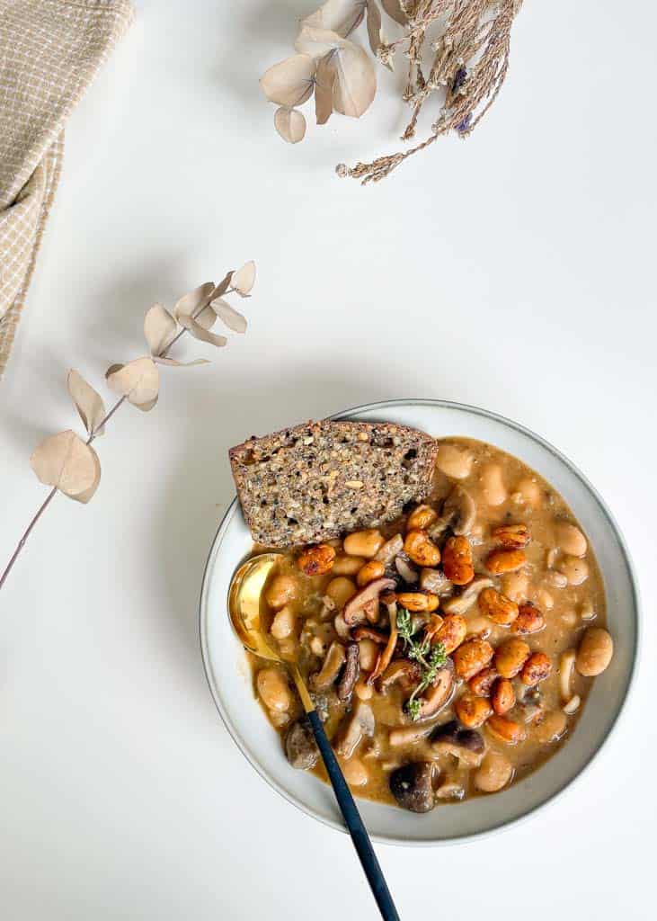 white bowl filled with white bean and mushroom soup topped with crispy white beans and fresh thyme with a piece of gluten free bread in the bowl and a gold and black spoon