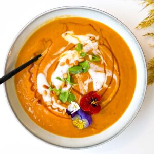 roasted butternut squash soup in a white bowl topped with a dollop of yogurt honey micro greens edible flowers and toasted pine nuts with a spoon in the bowl