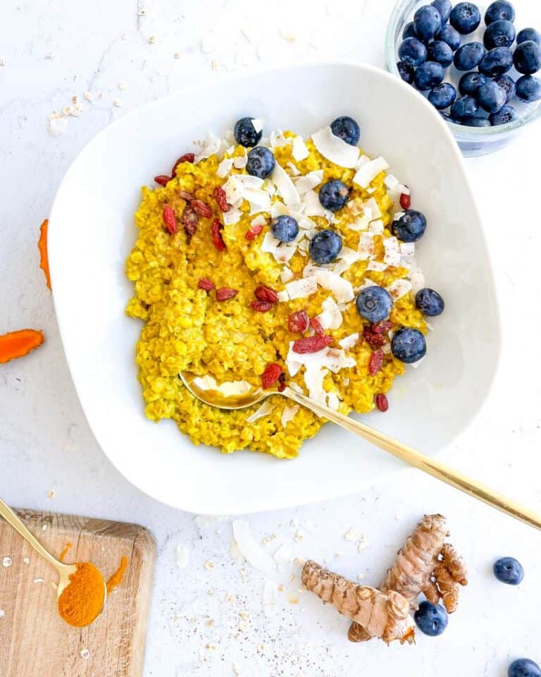 white bowl filled with turmeric oatmeal and topped with blueberries shredded coconut goji berries with a spoon in the bowl fresh turmeric blueberries are in the background