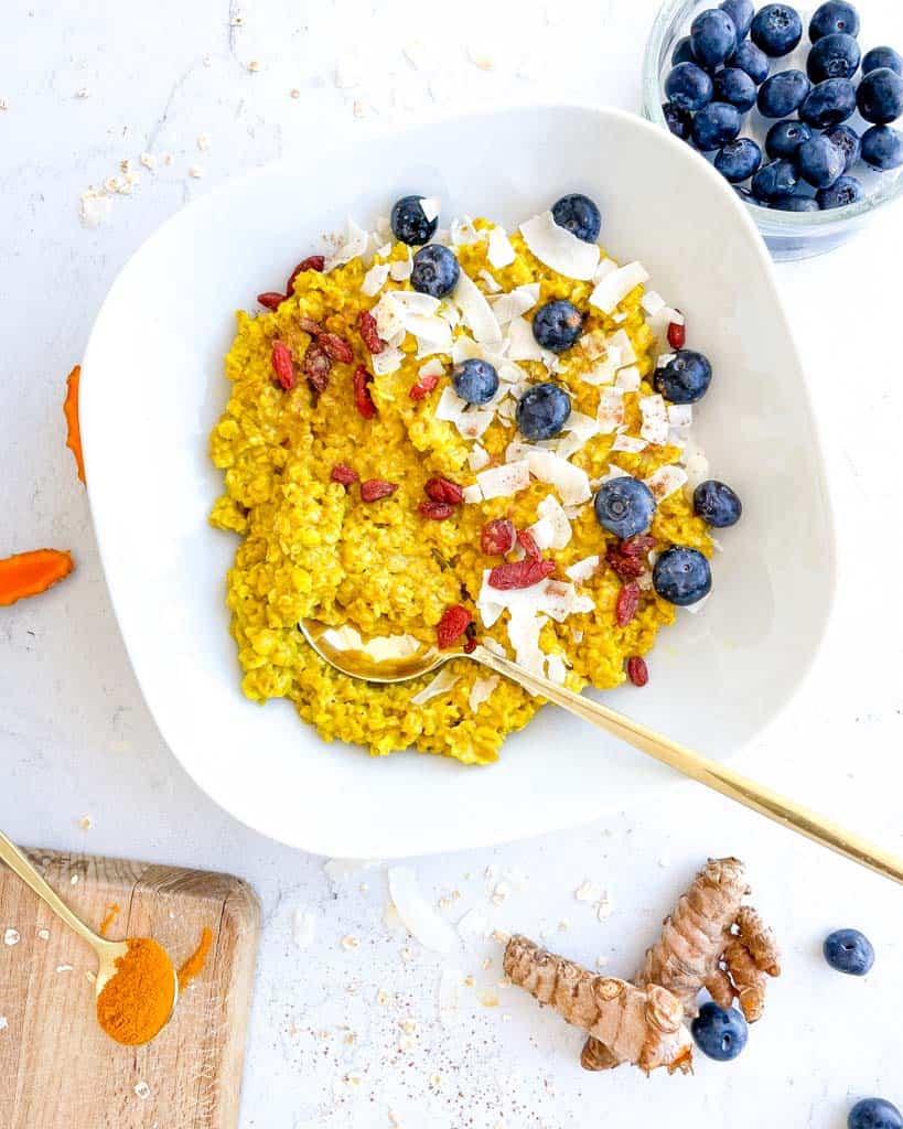 white bowl filled with turmeric oatmeal and topped with blueberries shredded coconut goji berries with a spoon in the bowl fresh turmeric blueberries are in the background